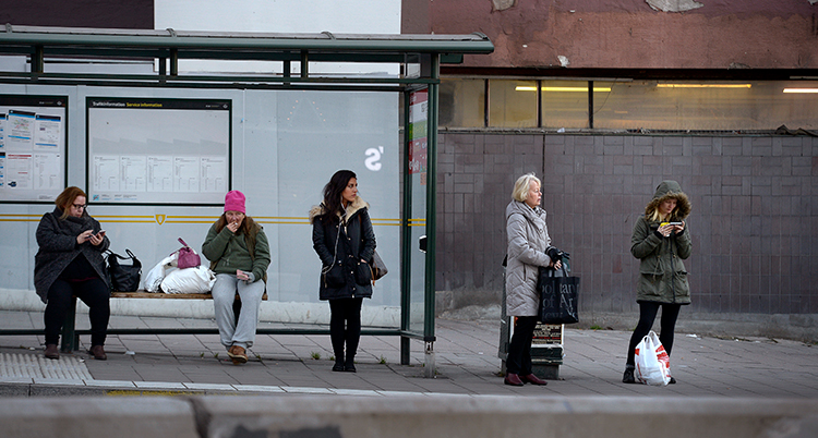 Folk väntar vid en busshållsplats. De står en bit ifrån varandra.