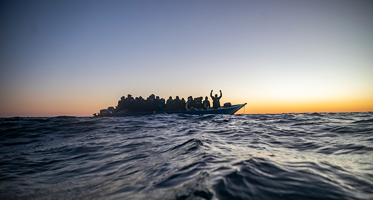 En båt full av människor syns på en stor havsvåg mot en himmel med en solnedgång.