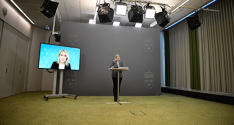 Regeringens utredare berättar om undersökningen. Hon står bakom ett litet bord i ett stort tomt rum. Till vänster om henne en stor tv-skärm med ministern Lena Hallengrens ansikte.