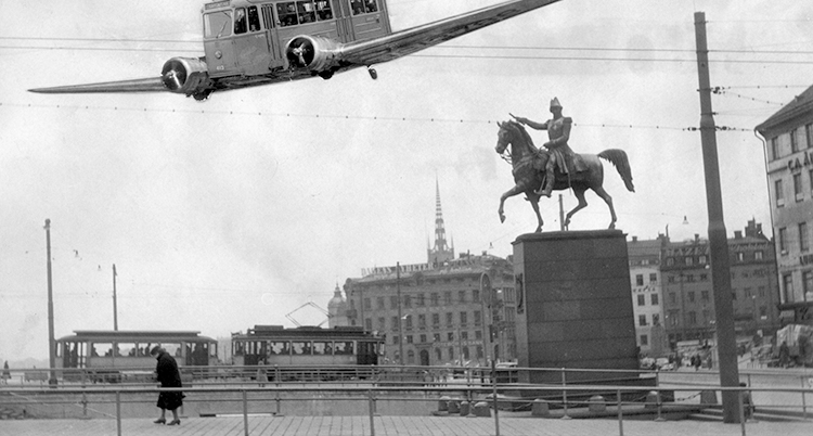 En farkost syns i himlen på en svartvit bild. Hus och spårvagnar syns som fond och en person går på ett torg under farkosten.