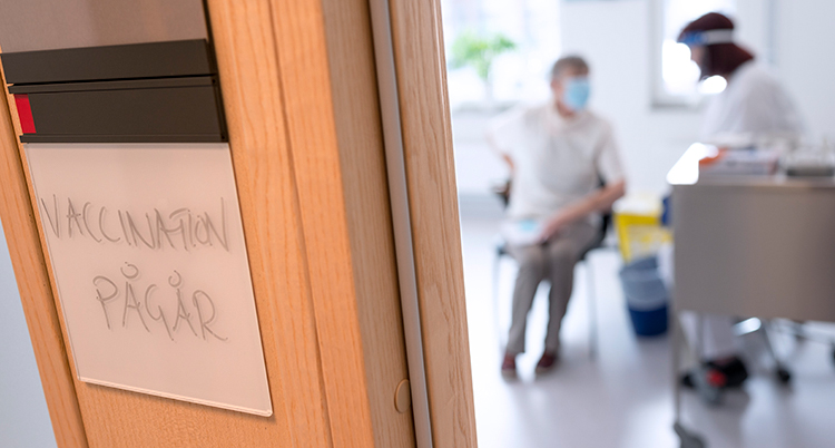 En bild på en dörr. Där ä en skylt med texten vaccination pågår. I bakgrunden syns en sköterska och en person som ska få en spruta.