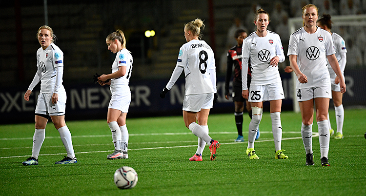 Några kvinnliga spelare går runt på en fotbollsplan. De ser inte glada ut. De har vita kläder. Matchen har precis slutat.