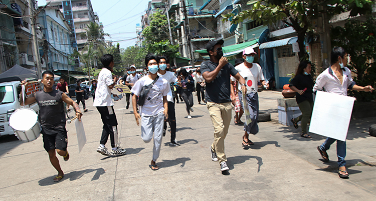 Bilden är från en stad i landet Myanmar. Människor springer på en gata. De springer mot kameran. Flera har munskydd på sig.