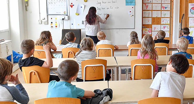 Bilden är tagen i ett klassrum i en skola. Vi ser ryggen på barnen. De sitter och lyssnar på läraren. Läraren står längst fram och skriver på tavlan. Barnen går i klass ett.