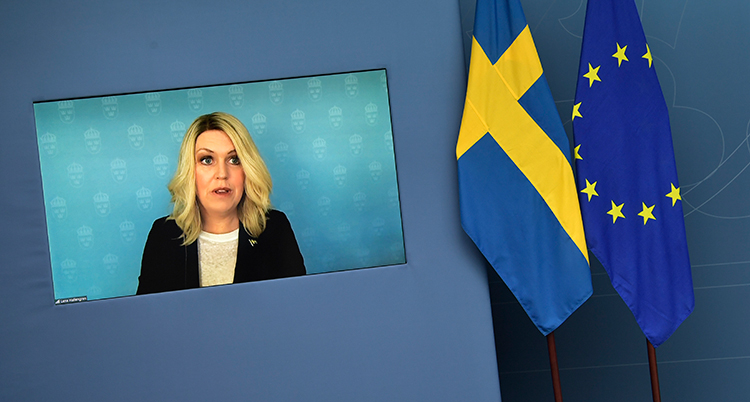 Lena Hallengren på en tv-skärm. Bredvid henne två flaggor, en svensk och EUs.