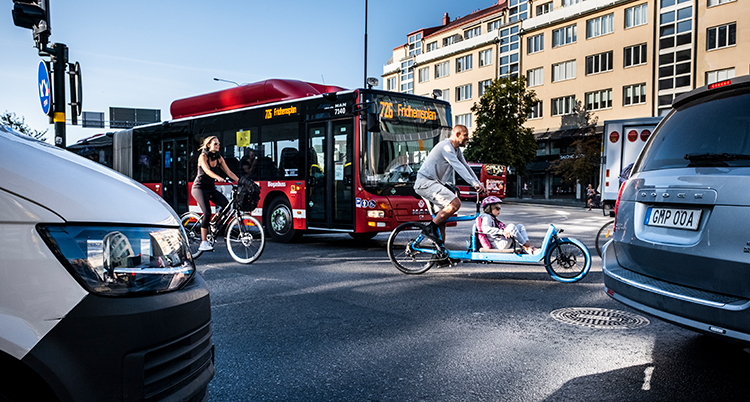 Trängsel på en gata. Ett par bilar är närmast kameran. Sedan kommert två cyklister framför en buss.