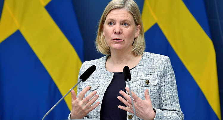 Magdalena Andersson har två mikrofoner framför sig. Hon pratar och gör en gest med händerna. Bakom henne är en svensk flagga.