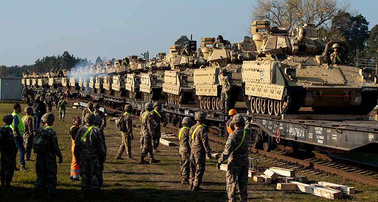 En lång rad med stridsvagnar som står på vagnar. Troligen är det vagnar till ett tåg. Soldater och andra står bredvid och tittar.