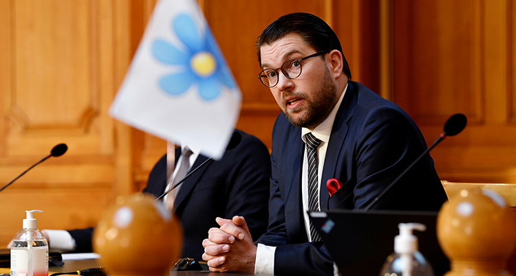 Åkesson sitter vid ett bord. Han ser allvarlig ut och har händerna knäppa på bordet framför sig. SDs flagga med en blåsippa står på bordet.