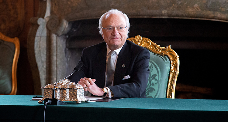 Kung Carl Gustaf sitter vid ett bord. Han har glasögon och kostym. Bordet har en grön duk. Fåtöljen som kungen sitter i är grön. I bakgrunden syns en öppen spis.