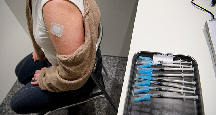 En person på en stol har ett plåster på armen efter vaccinering. Bakom på ett bord ligger flera nålar för vaccinering.