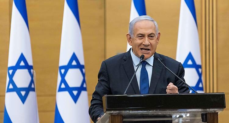 Netanyahu står och pratar i riksdagen. Han står i en talarstol och pratar i en mikrofon. Han har grått hår och kostym. I bakgrunden finns Israels flagga.