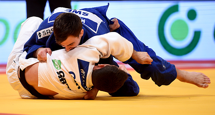 Från en match i judo. Två personer brottas på en matta. En person har blå dräkt och den andra har vit dräkt.