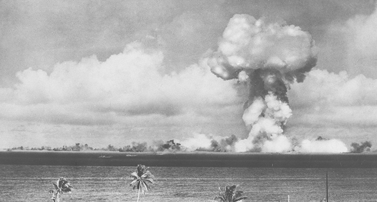 En svartvit bild över ett hav med palmer i förgrunden. Ett gigantiskt svampformat moln syns i horisonten.