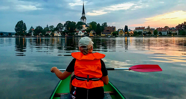 Barnet har en orange flytväst, sitter med ryggen mot kameran i en grön kanot. I horisonten syns en stad med en hög vit kyrka. Vattnet är blankt.