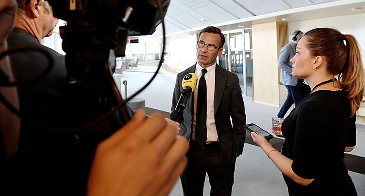 Ulf Kristersson står i riksdagens hus. Han pratar med journalister som har kameror och mikrofoner.