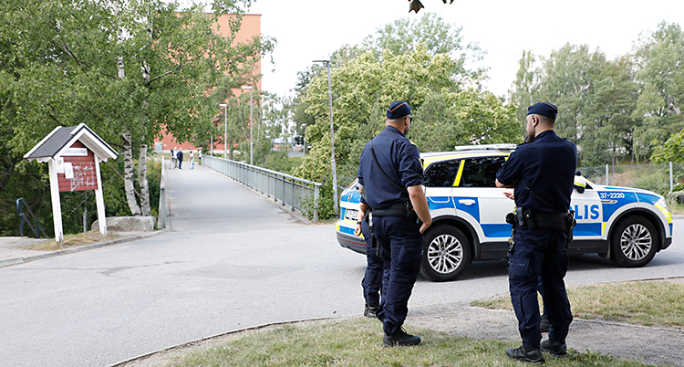 Bilden är tagen i Flemingsberg. Två poliser står utomhus, med ryggen mot kameran. De står vid en polisbil. Längre fram syns en gångbro.