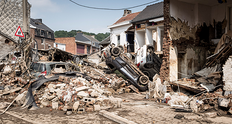 I en stad. Flera hus har blivit förstörda. Högarna efter husen ligger på marken. Där syns också tre förstörda bilar. Två av bilarna ligger upp och ner.