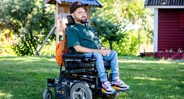 Han sitter i en rullstol som går på el. Han tittar in i kameran. Han har en keps och en grön t-shirt.