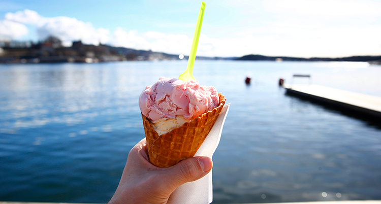 En hand håller i en glasstrut med vit och rosa glass i. Bakom syns vatten.