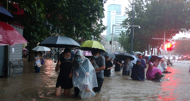 Människor går med regnkläder och paraplyer på en översvämmad gata. De har vatten långt upp på benen.