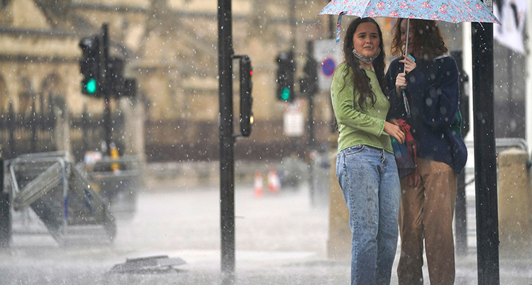 Två kvinnor står under ett paraply. Det regnar och är mycket vatten på gatan.