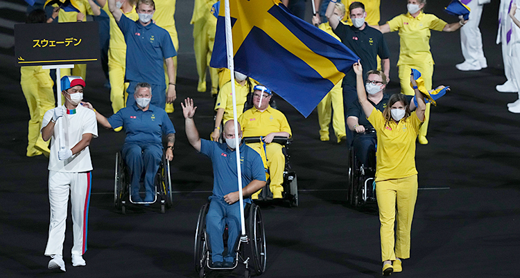 Svenska idrottare går in på en stor arena. De har gula och blå kläder. Några åker i rullstol.