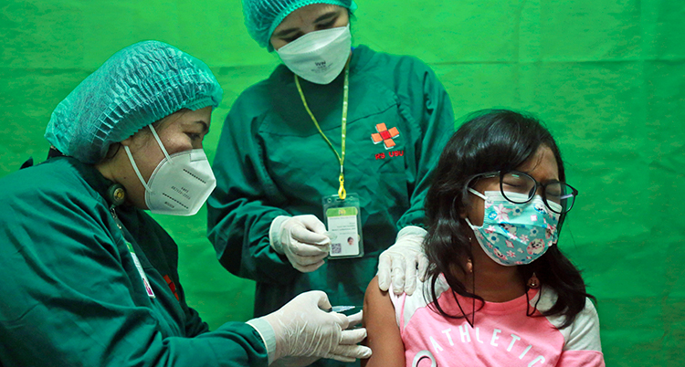 Två sjukvårdsklädda personer med munskydd och hätta ger en flicka i rosa tröja en spruta i armen. Flickan har munskydd och blundar med ögonen.