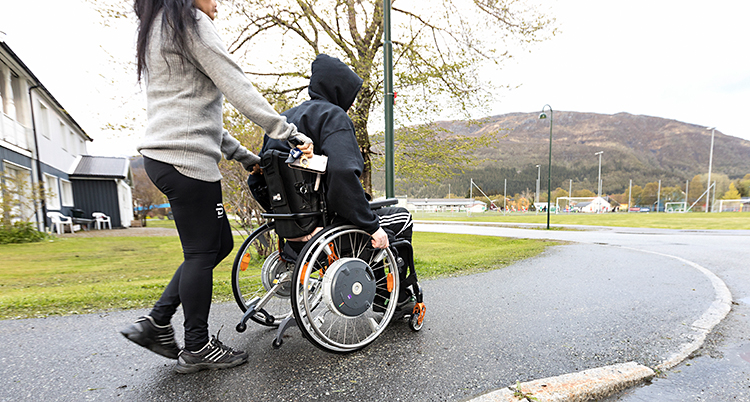 Bilden är tagen på en trottoar, utomhus. En person sitter i en rullstol. Bakom står en annan person och håller i rullstolen.