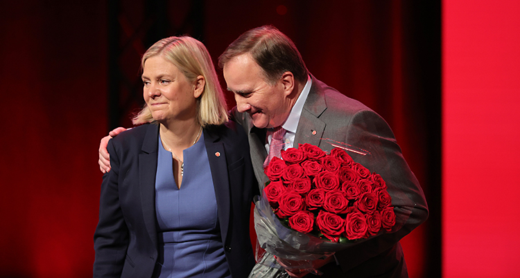 De står på en scen. Stefan Löfven har en bukett med röda rosor i handen.