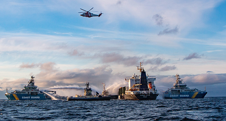 En bild från havet där flera båtar står runt om ett stort fartyg. En helikopter är i luften.