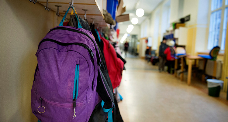 en ryggsäck i förgrunden hänger på en krok i en skolkorridor.