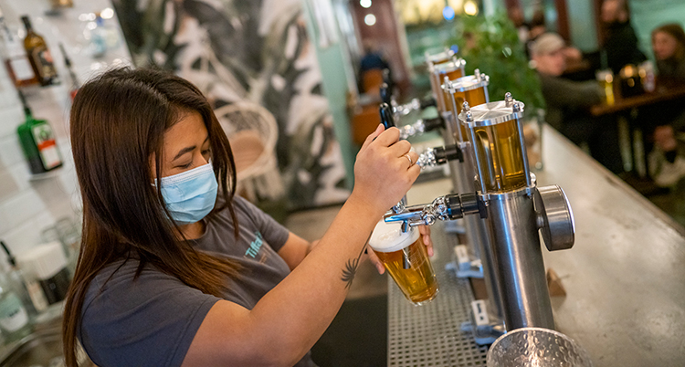 En bartender häller upp en öl i en bar. Hon har munskydd på si.