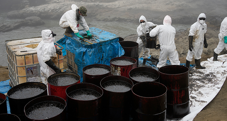 Människor i skyddande dräkter jobbar med att ta bort olja vid stora tunnor för olja.
