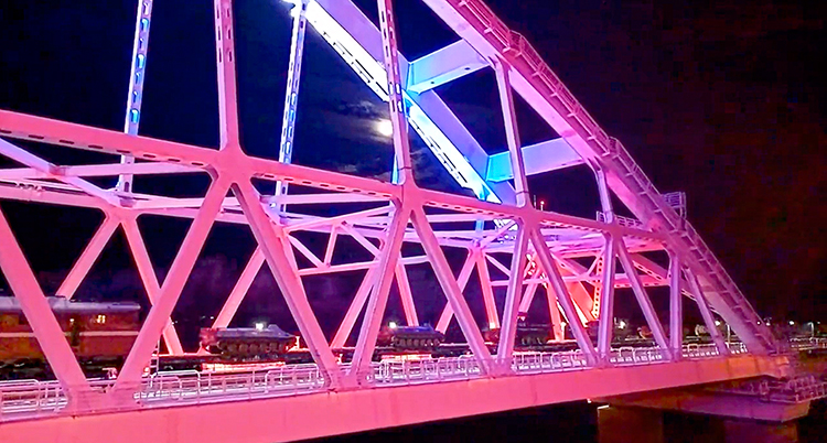 En bro upplyst i rosa ljus mot en svart himmel. Fordon kör på bron.