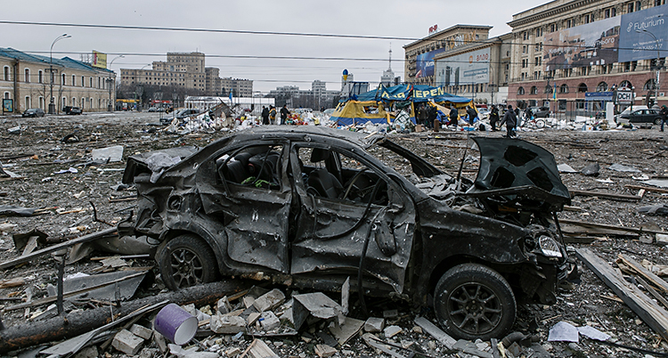Ett torg i staden Charkiv. En bil är helt svart och förstörd. Det ligger skräp överallt.