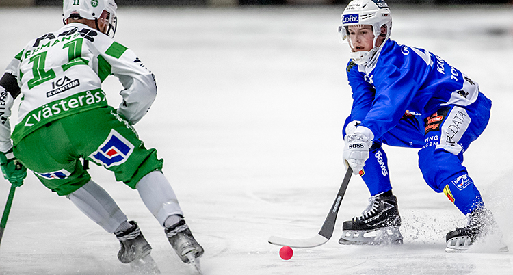 Från en match i bandy. En spelare med blå dräkt har bollen. En spelare med grön dräkt jagar efter.