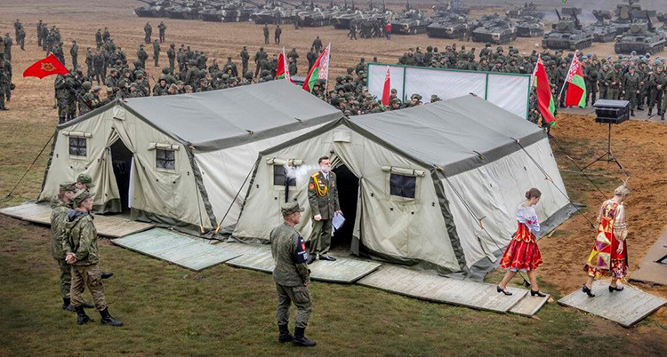 En militär står framför militärtält och röker. Två kvinnor går bort från tälten. Bakom tälten finns många militärer. De är på en övningsplats på ett fält.
