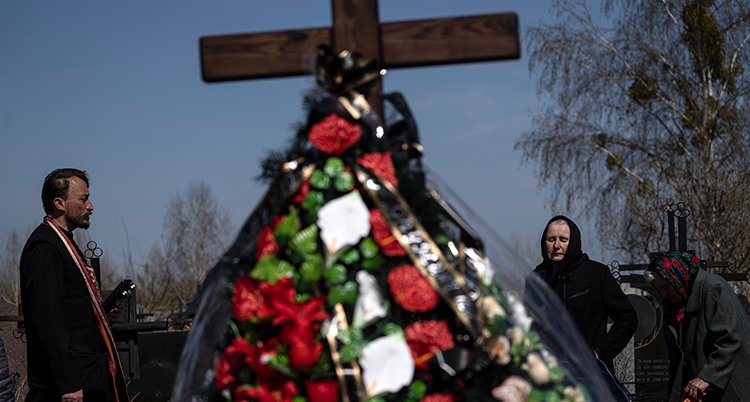 En man står i profil till vänster i bild. Han tittar mot ett kors. Under korset finns många blommor. Till höger om korset står en ledsen kvinna med en sjal på huvudet.