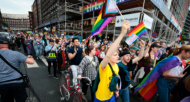 Många människor går på en gata i en stad. De demonstrerar. En del har regnbågsflaggor i händerna och sträcker flaggorna upp i luften.