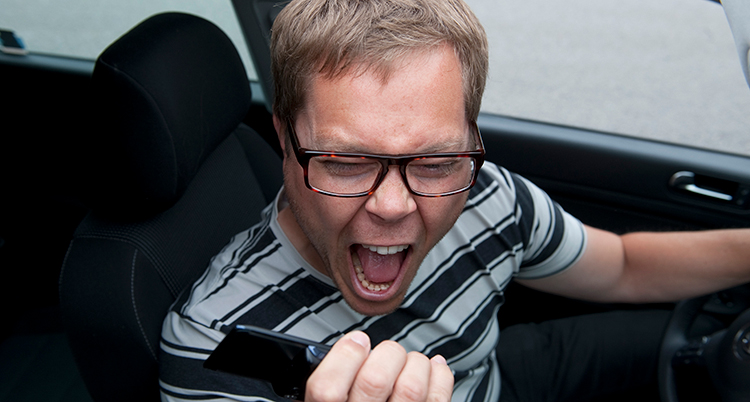 En arg man skriker in i en mobil. Han sitter vid ratten i en bil.