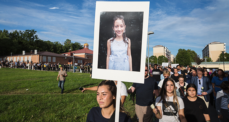 En tjej syns längst fram i ett långt demonstrationståg. Hon håller upp en skylt med en bild av en flicka. Demonstrationståget går över en gräsplan. Bakom syns hyreshus. Himlen är blå.
