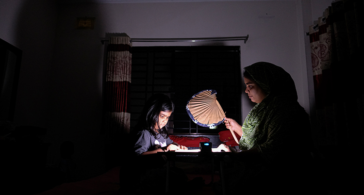 Ett mörkt rum med en flicka och en kvinna som sitter vid ett bord. Det är en liten lampa som lyser vid bordet.