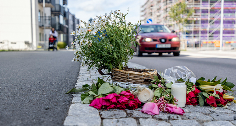 Blommor och ljus ligger på trottoaren. I bakgrunden syns en bil och hus.