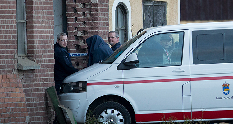En vit skåpbil är parkerad med fronten mot ett hus. Flera vårdare tittar mot kameran, samtidigt som en man med svart jacka över huvudet blir förd från bilen mot byggnaden.