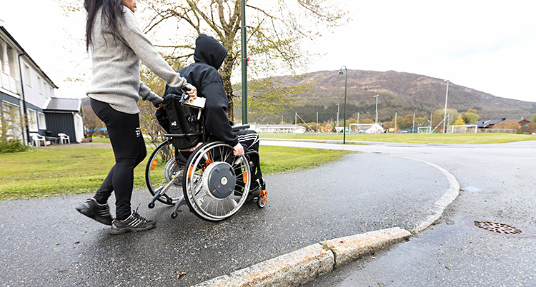 En kvinna skjutsar en man som sitter i en rullstol. De går på en trottoar.