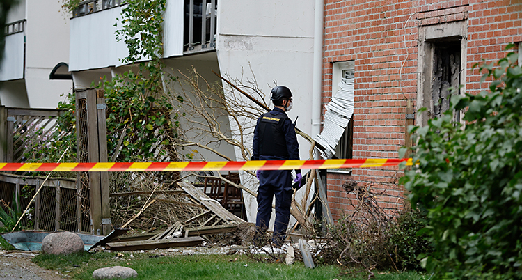 En polis står utanför ett hus med lägenheter. Flera fönster är förstörda.