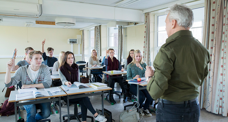 Bilden är från ett klassrum. En lärare har en lektion. Läraren är en man. I klassrummet sitter elever i bänkar.