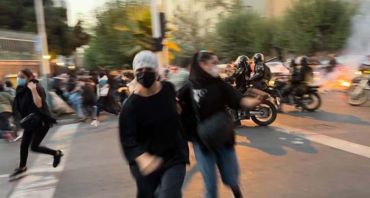 några kvinnor med masker för ansiktet springer iväg från en gata. Något brinner längre bort på gatan. Det syns också en folksamling och personer på motorcyklar längre bort på gatan.