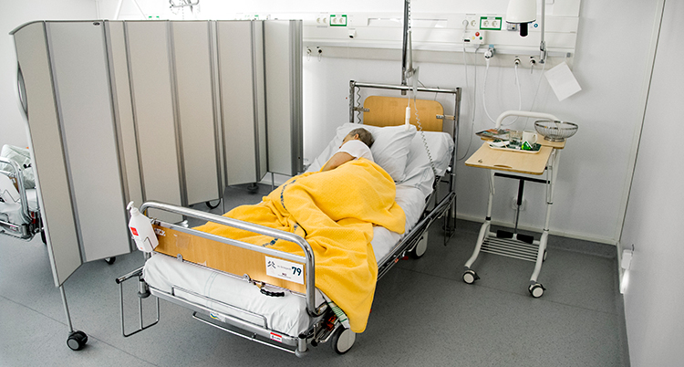 En person ligger under en filt i en säng i ett rum på ett sjukhus